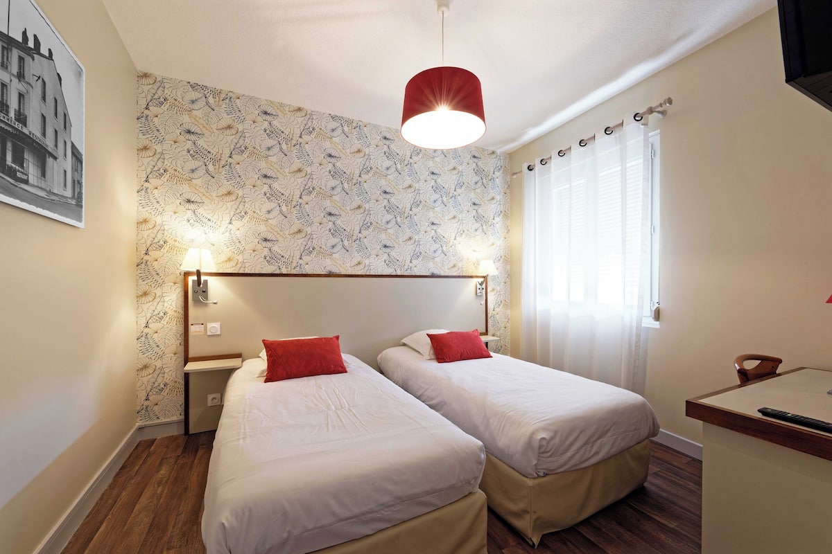 Une chambre d’hôtel avec deux lits, un plancher en bois, un papier peint à motifs, un luminaire suspendu rouge et une fenêtre avec des rideaux.