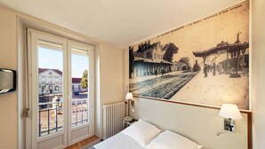 Una habitación de hotel con una cama, un ventanal con vista a un edificio y una lámpara fijada a la pared.