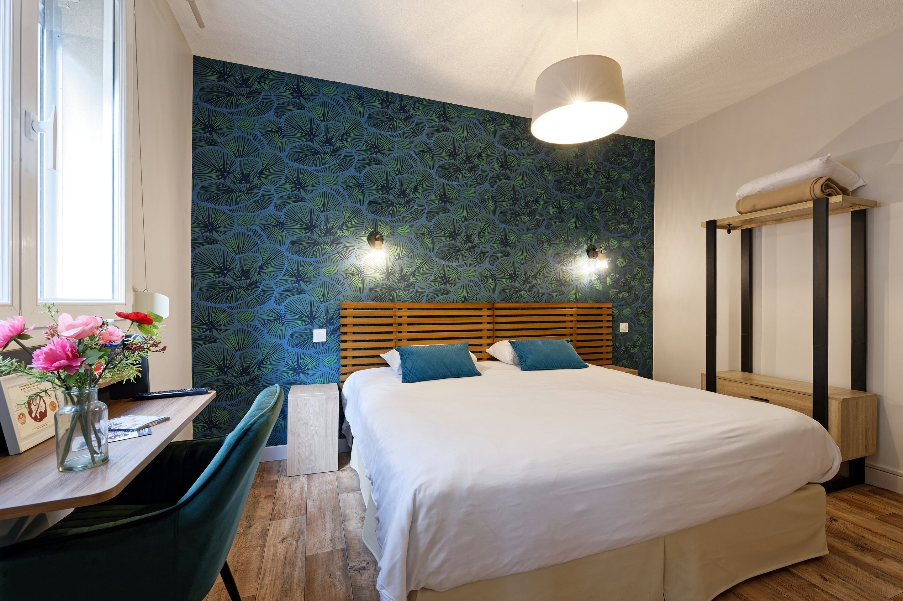 Une chambre à coucher comprenant un lit, des tables de chevet, un bureau avec une chaise, une fenêtre et un mur orné d’un motif de feuilles.