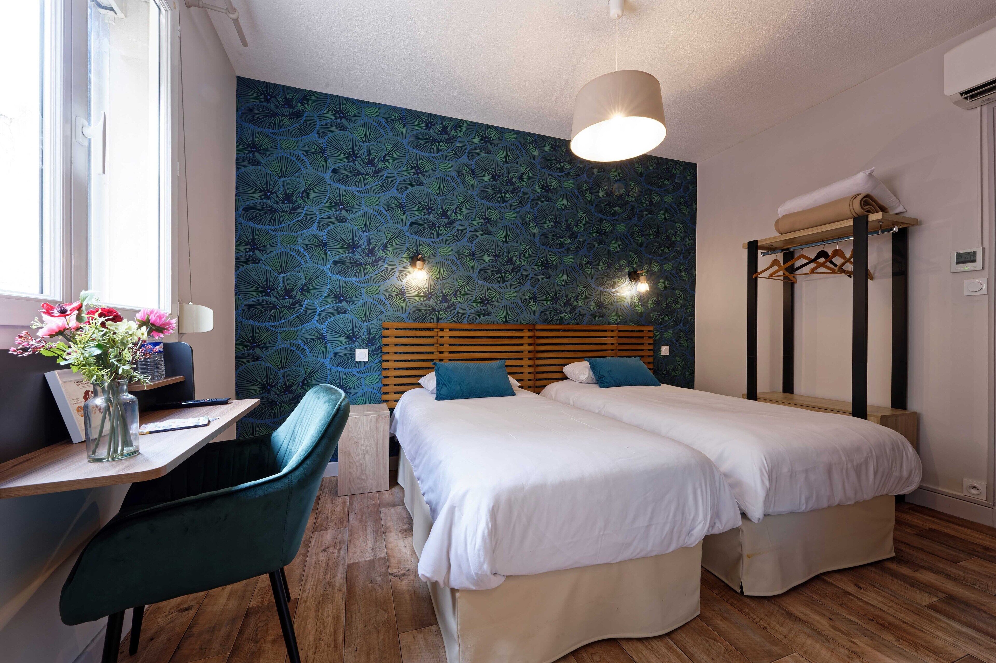 Habitación de hotel con dos camas, un escritorio y una silla, cabecera de madera, pared con diseño azul y una lámpara colgante.