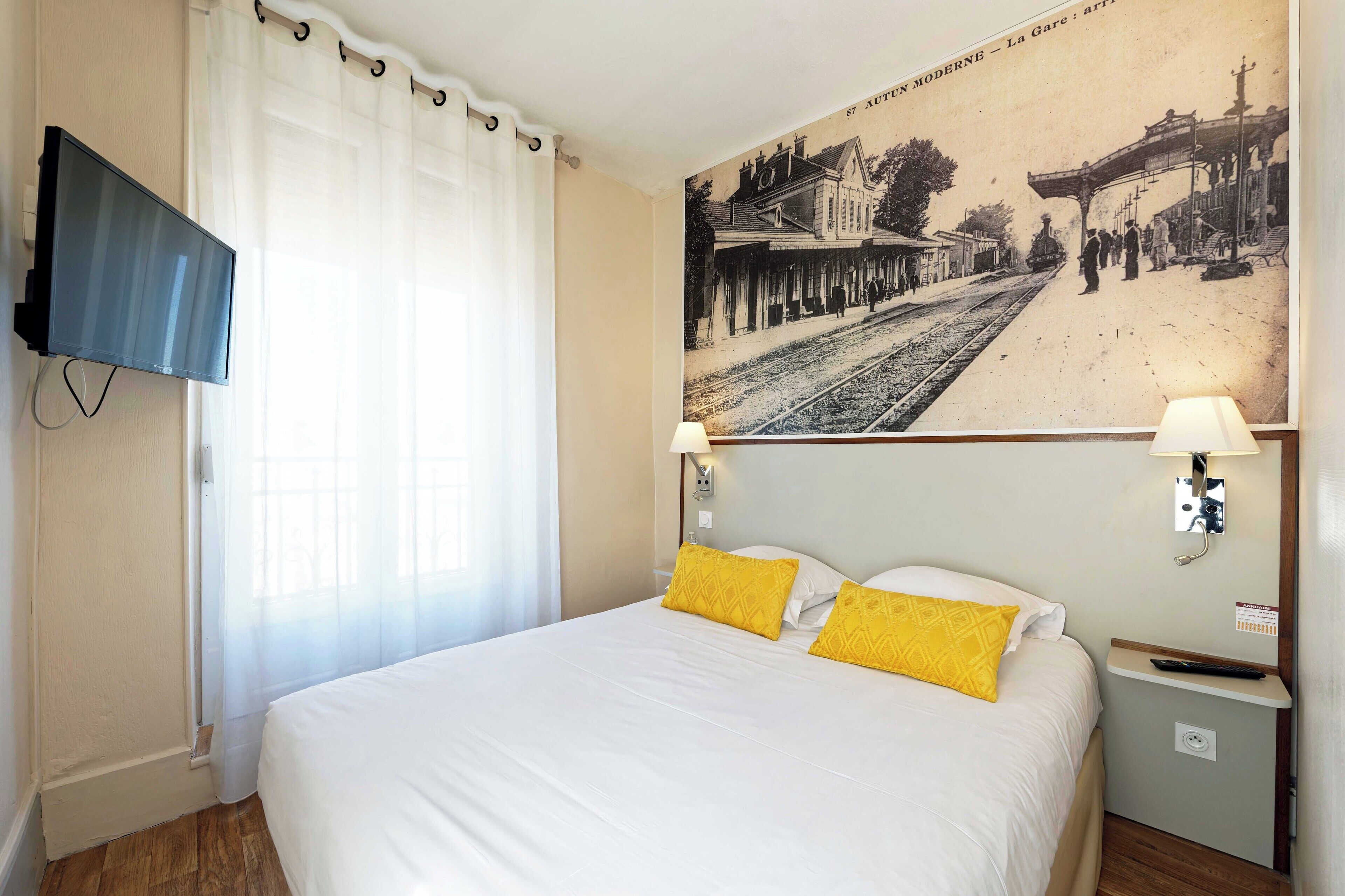 Una habitación de hotel con una cama, un televisor montado en la pared, un cuadro de una estación de tren y una mesita de noche con una lámpara.