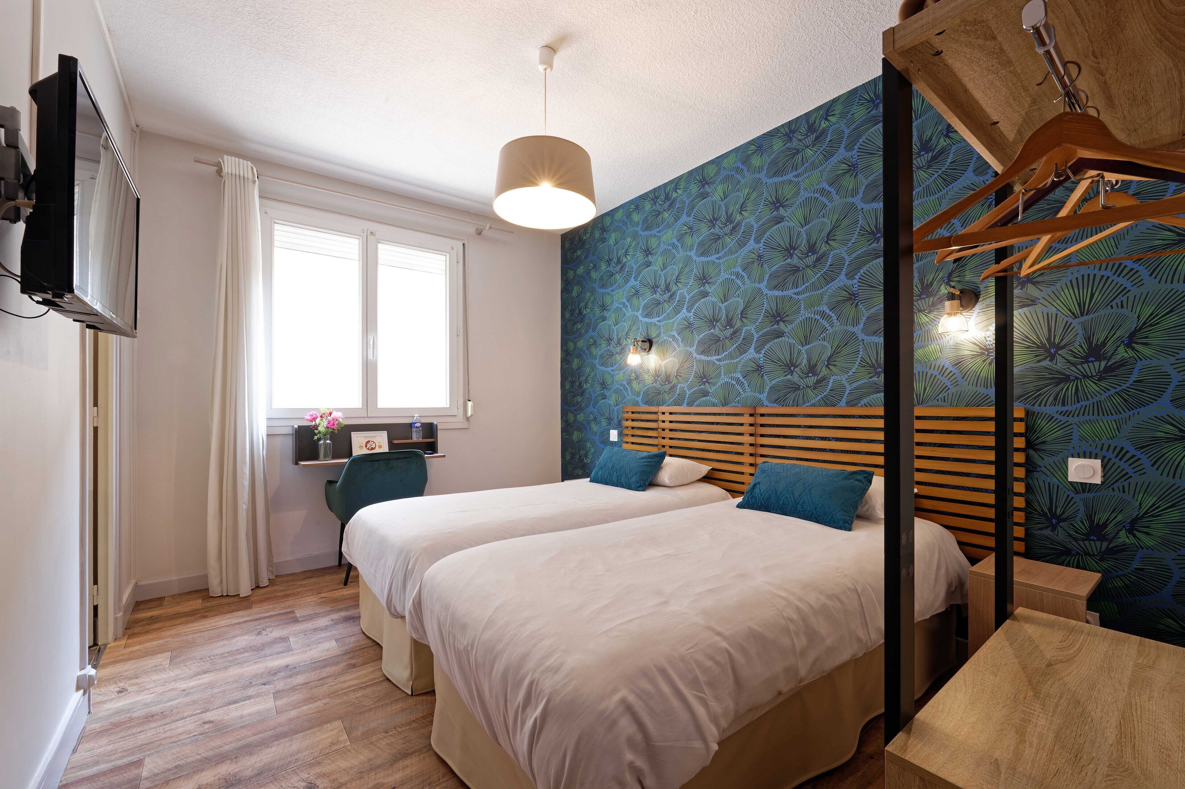Habitación de hotel con dos camas, cabecero de madera, televisor, ventana con cortinas y pared con un diseño de hojas azules.