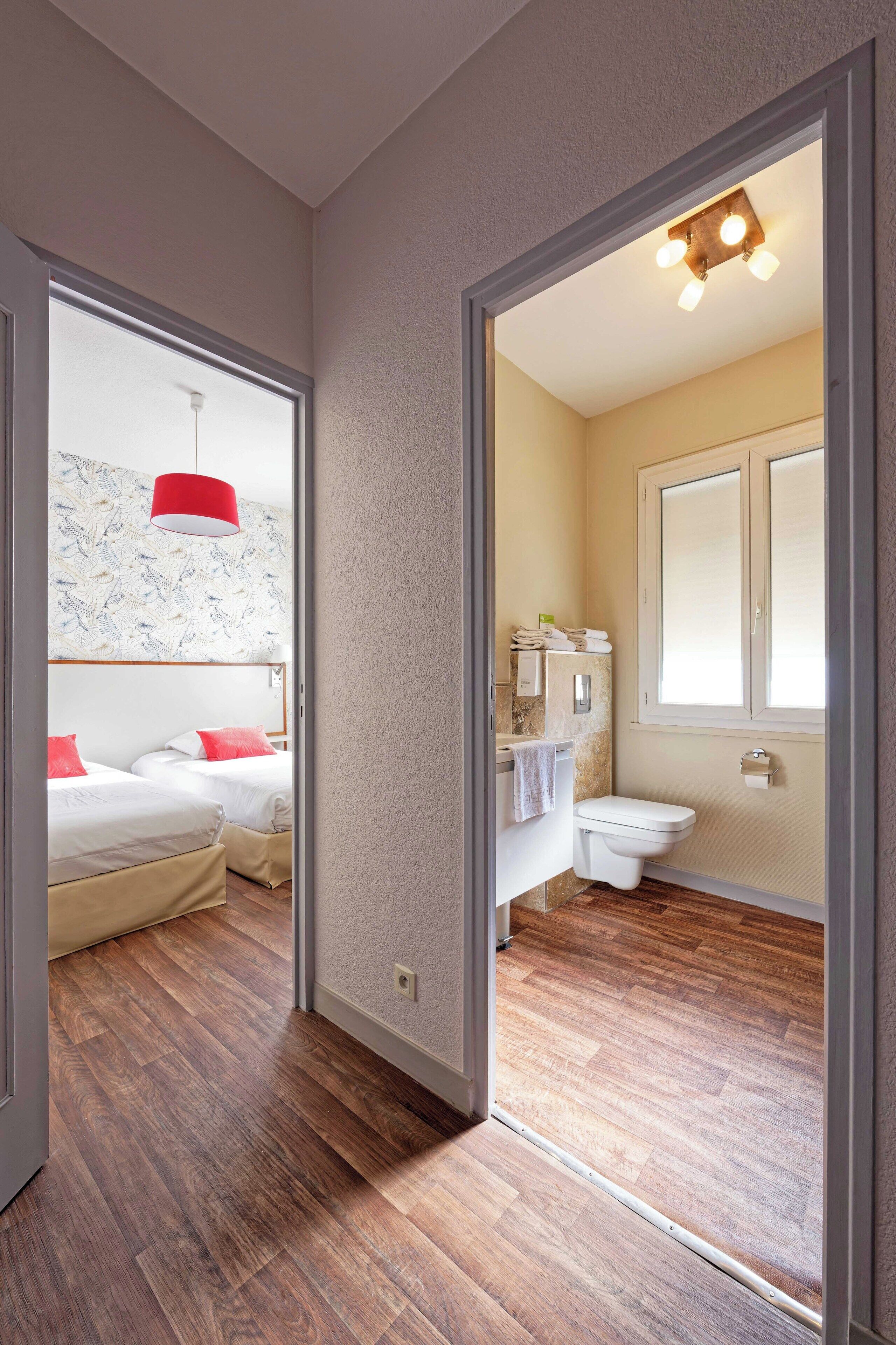 A hotel room with a wooden floor, a red pendant light, a bed with white bedding and red pillows, a bathroom with a white toilet and sink, and a window with a view.