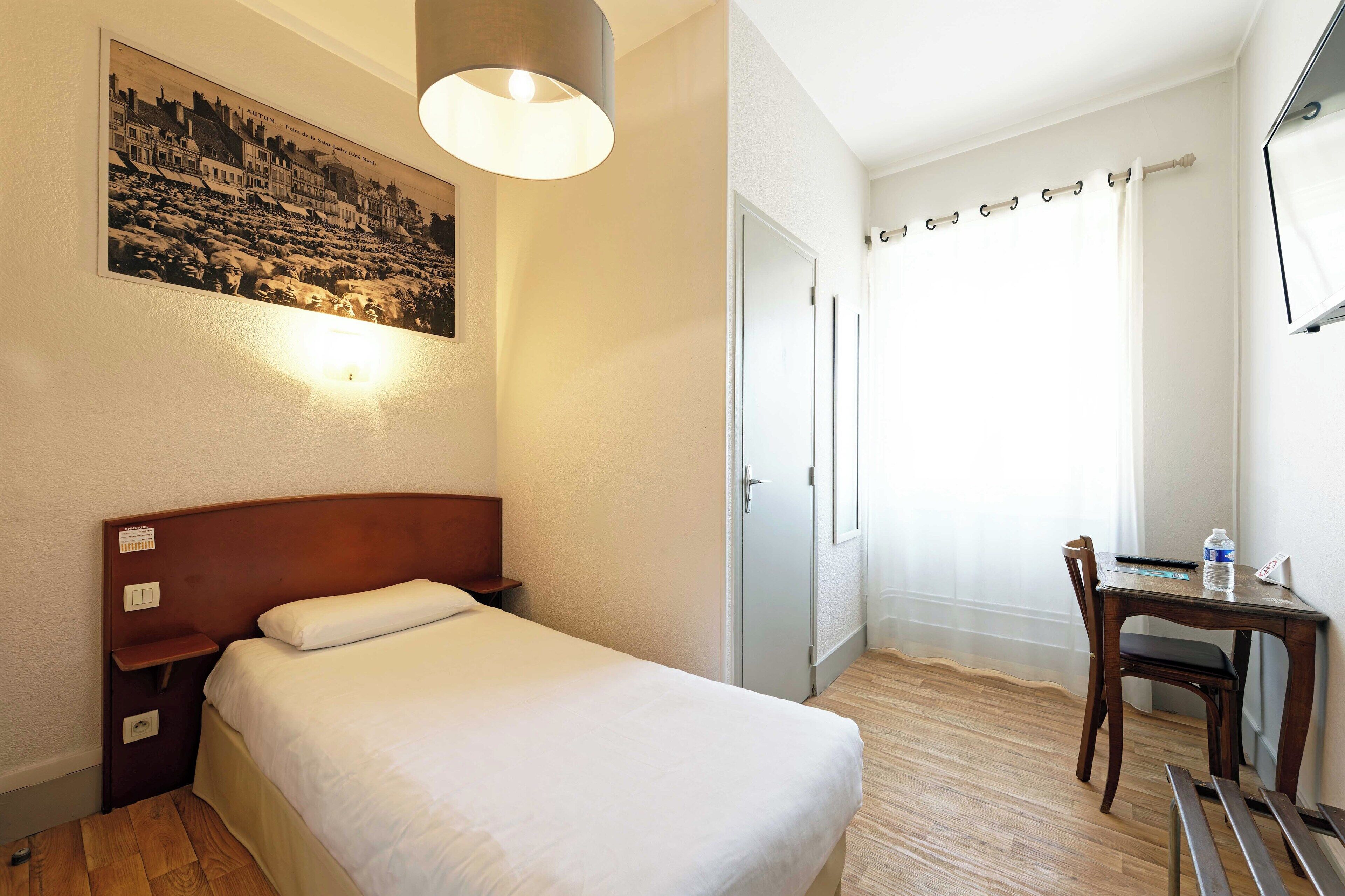 A hotel room with a bed, bedside table, lamp, desk with chair, and a painting on the wall.