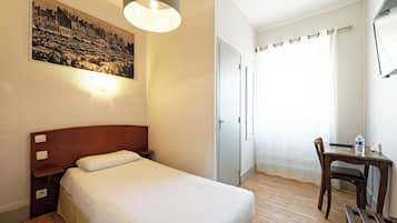 A hotel room with a bed, bedside table, lamp, desk with chair, and a painting on the wall.