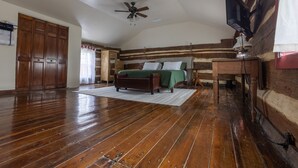 Une chambre avec un plancher en bois, un lit recouvert d’une couverture verte, un ventilateur de plafond et un téléviseur fixé au mur.