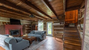 L'intérieur d'une cabane en rondins, avec une cheminée, un canapé, un téléviseur et un escalier menant à un espace extérieur.