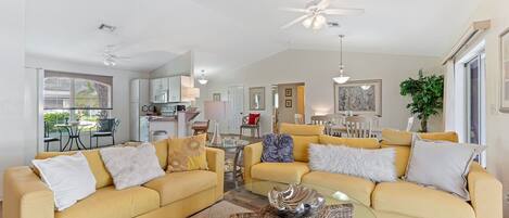 A spacious living room with two yellow sofas, a glass coffee table, and a ceiling fan.