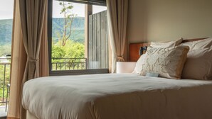 Een slaapkamer met een groot bed, beige gordijnen, een nachtkastje en uitzicht op een bergachtig landschap.