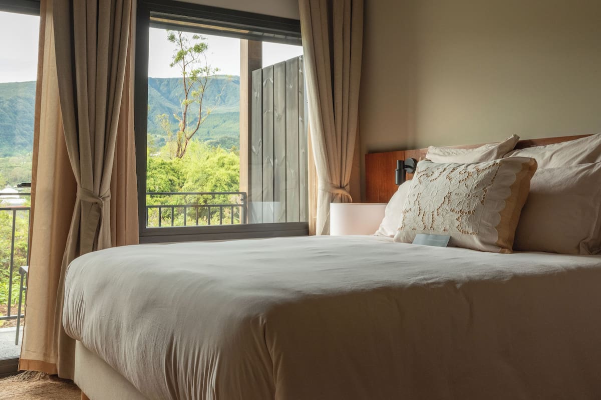 Un dormitorio con una cama grande, cortinas color beige, una mesita de noche y vistas a un paisaje montañoso.