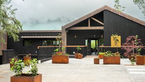 Een modern gebouw met een donkere gevel, grote ramen en een schuin dak. Voor de ingang staan meerdere plantenbakken met groen.