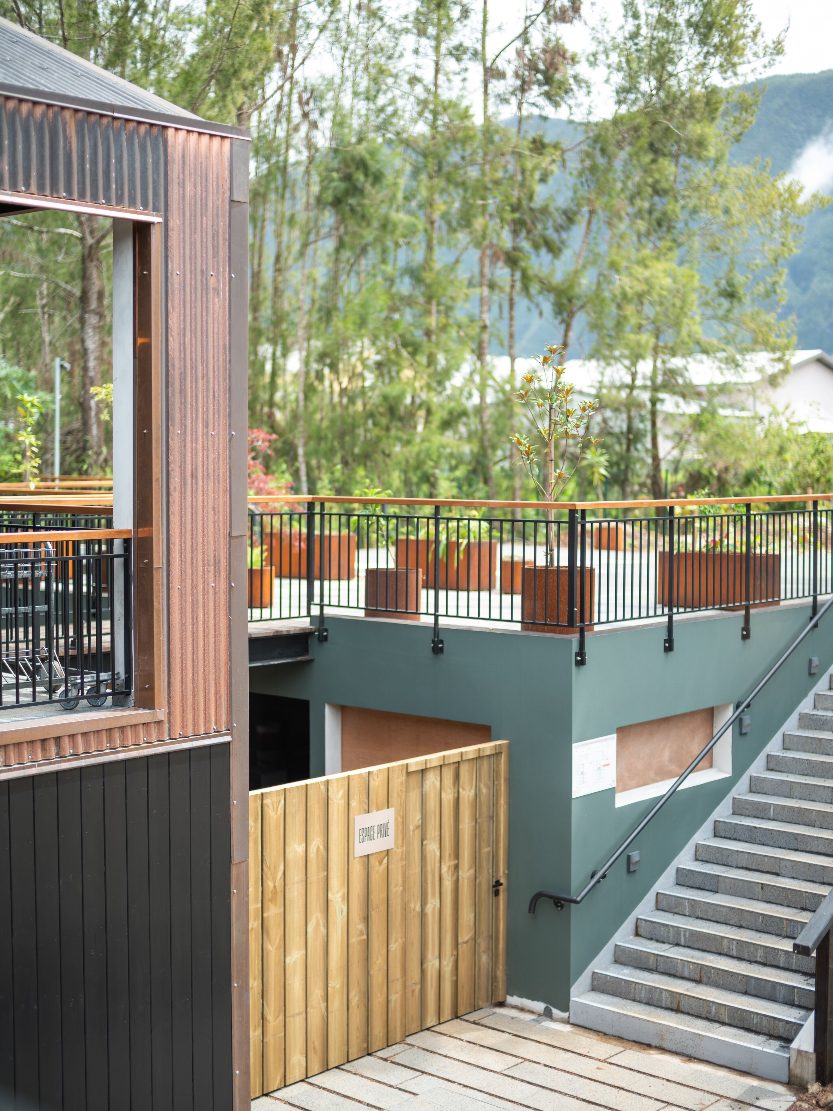 A wooden cabin with a staircase leading to a deck, surrounded by greenery and mountains.