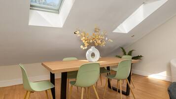 A dining area with a wooden table, four green chairs, a white vase with dried flowers, and a skylight.