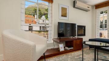 A living room with a white armchair, a wooden entertainment unit with a TV, and a view of a hillside through a large window.