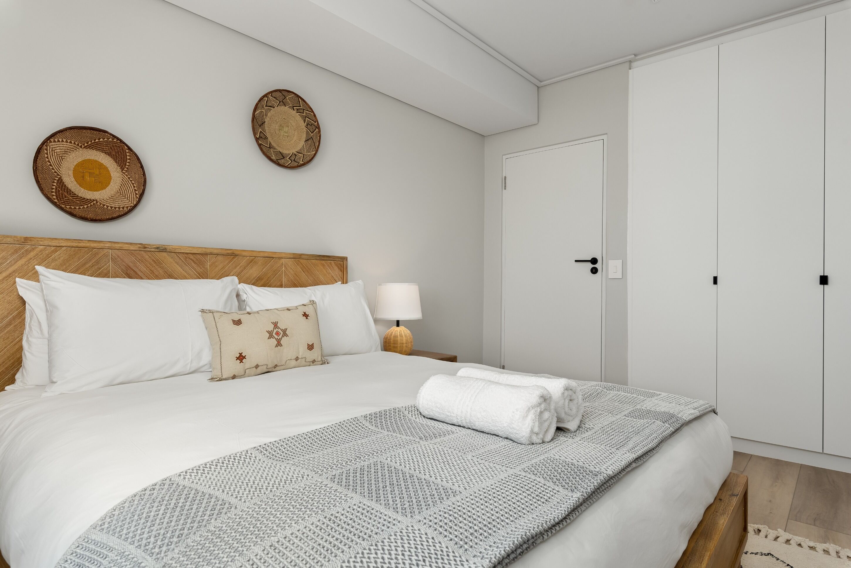 A neatly made bed with white linens and a patterned throw, a wooden headboard, and two wall-mounted woven decorations.