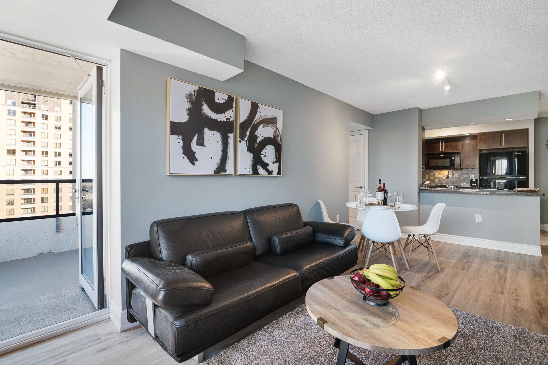 A modern living room with a black leather sofa, a round wooden coffee table with fruit, and two white chairs. There are abstract paintings on the wall, a kitchen area with dark cabinets, and a view of a building through a glass door.