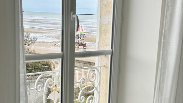 Una mesa con una botella de champán y copas, una ventana con vista a una playa y un tractor rojo, y una cortina blanca.