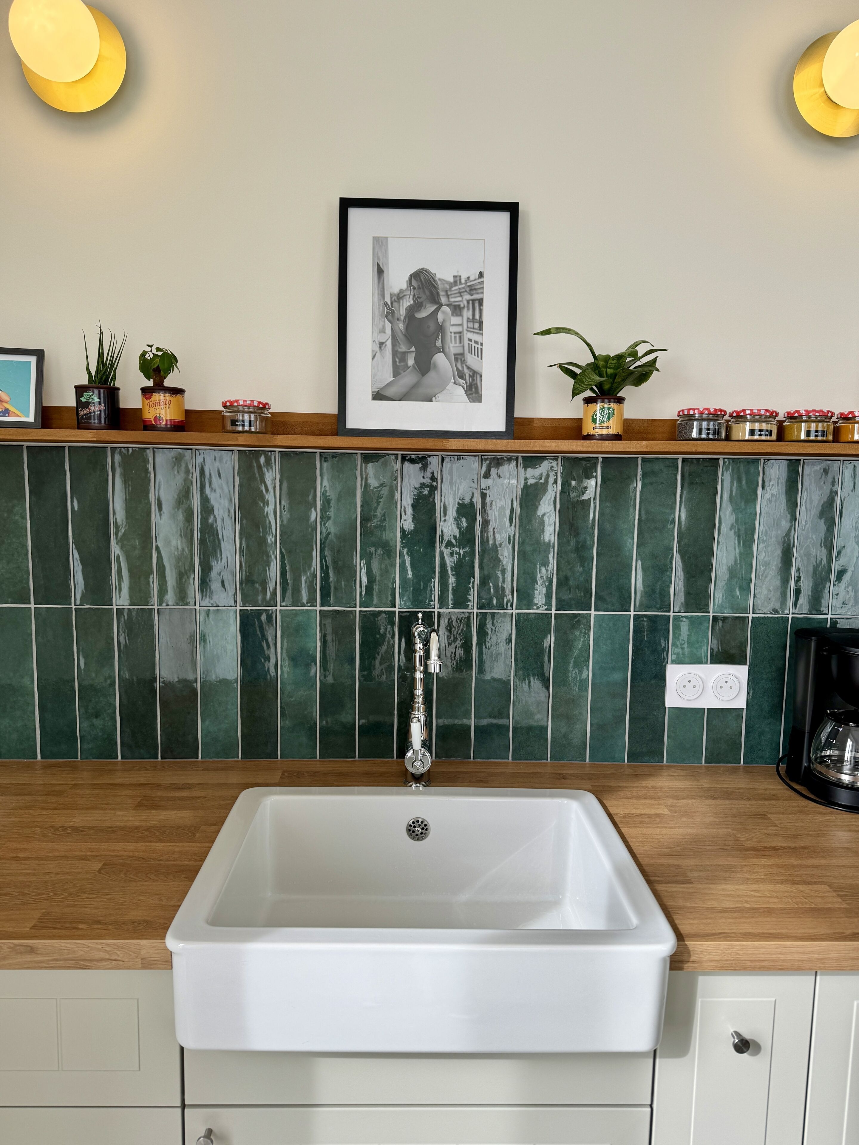 Cocina con pileta blanca, encimera de madera, salpicadero de azulejos verdes y un cuadro colgado sobre la pileta.