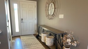 A hallway with a wooden floor, a patterned rug, a bench, a vase with dried flowers, and a door with a mirror above it.