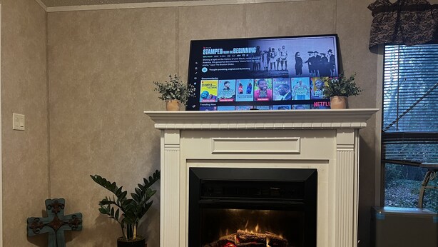 Un salon avec une cheminée, un téléviseur fixé au-dessus diffusant une émission de Netflix, et une plante en pot placée à gauche.
