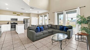 A spacious living area with a gray sectional sofa, a glass coffee table, and a kitchen with white cabinets and stainless steel appliances. - EV_8583 - 8 Bed 4.5 Bath Villa with Screened-In Private Pool and Access to Resor (Kissimmee)