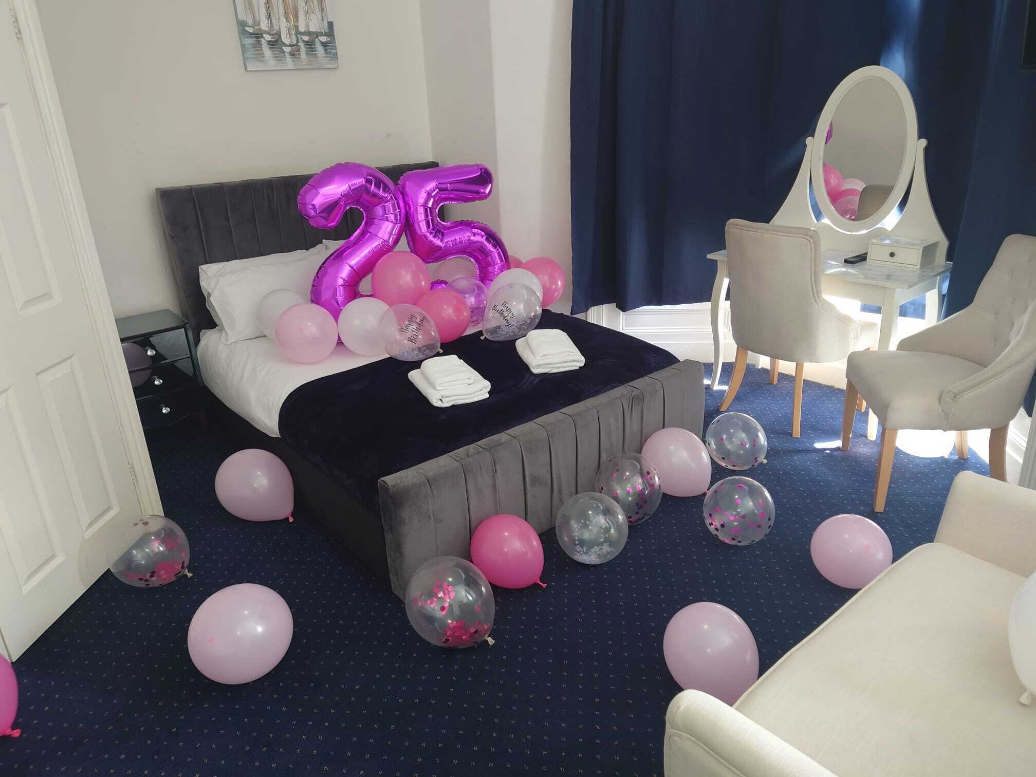A hotel room with a bed, a bedside table, a chair, and a mirror. There are balloons scattered around the room.