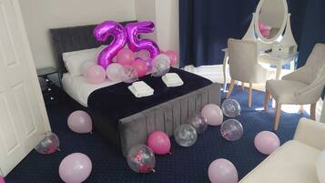 A hotel room with a bed, a bedside table, a chair, and a mirror. There are balloons scattered around the room.