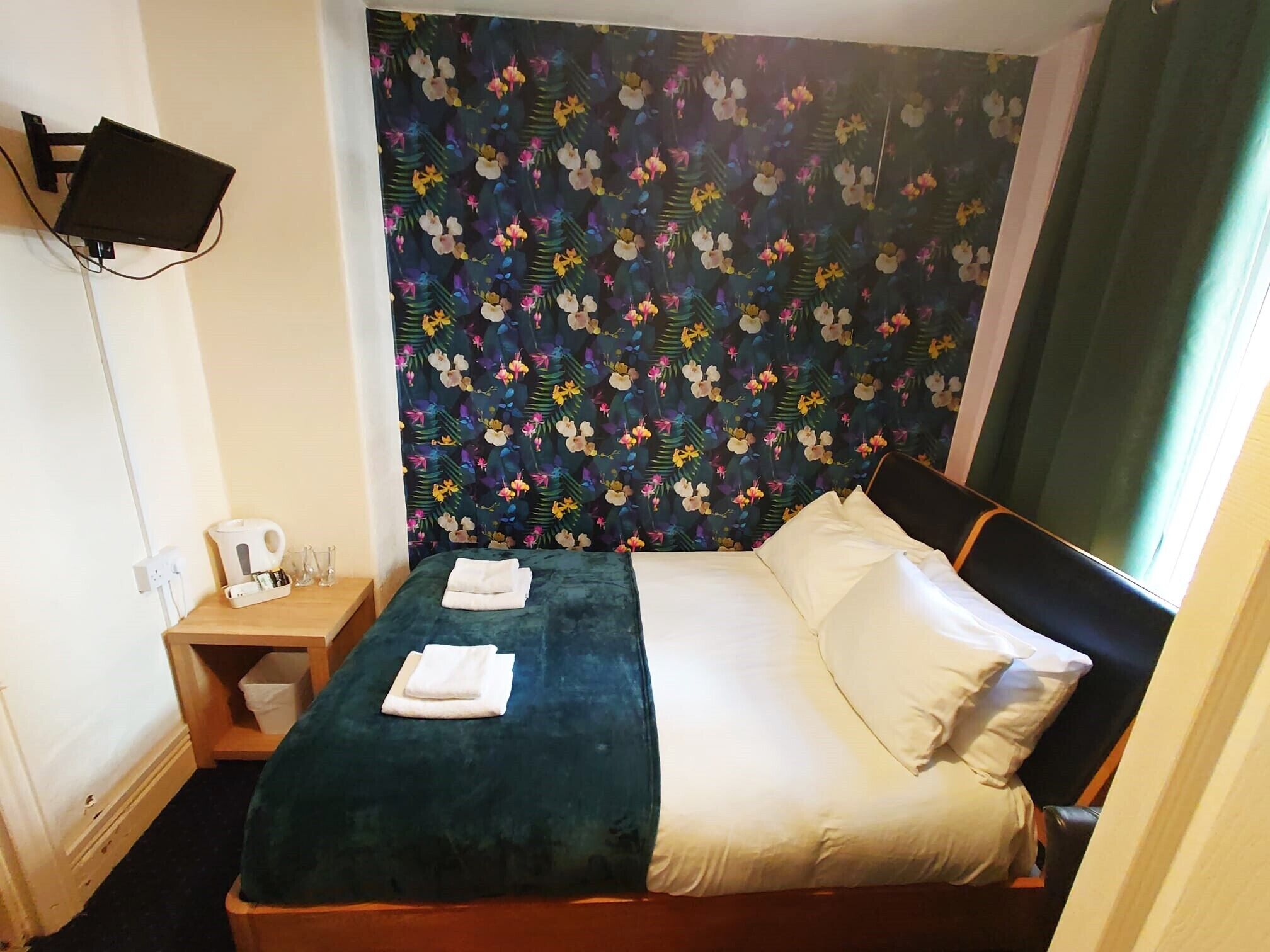 Habitación de hotel con cama, mesita de noche, hervidor y cabecera con estampado floral.