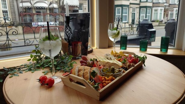 A table with a charcuterie board, glasses of water, and a view of a city street through a window. - Harriette's Guest House (Blackpool)