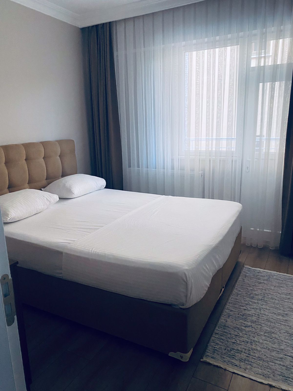 Un dormitorio con una cama grande, cabecera y una ventana con cortinas traslúcidas.