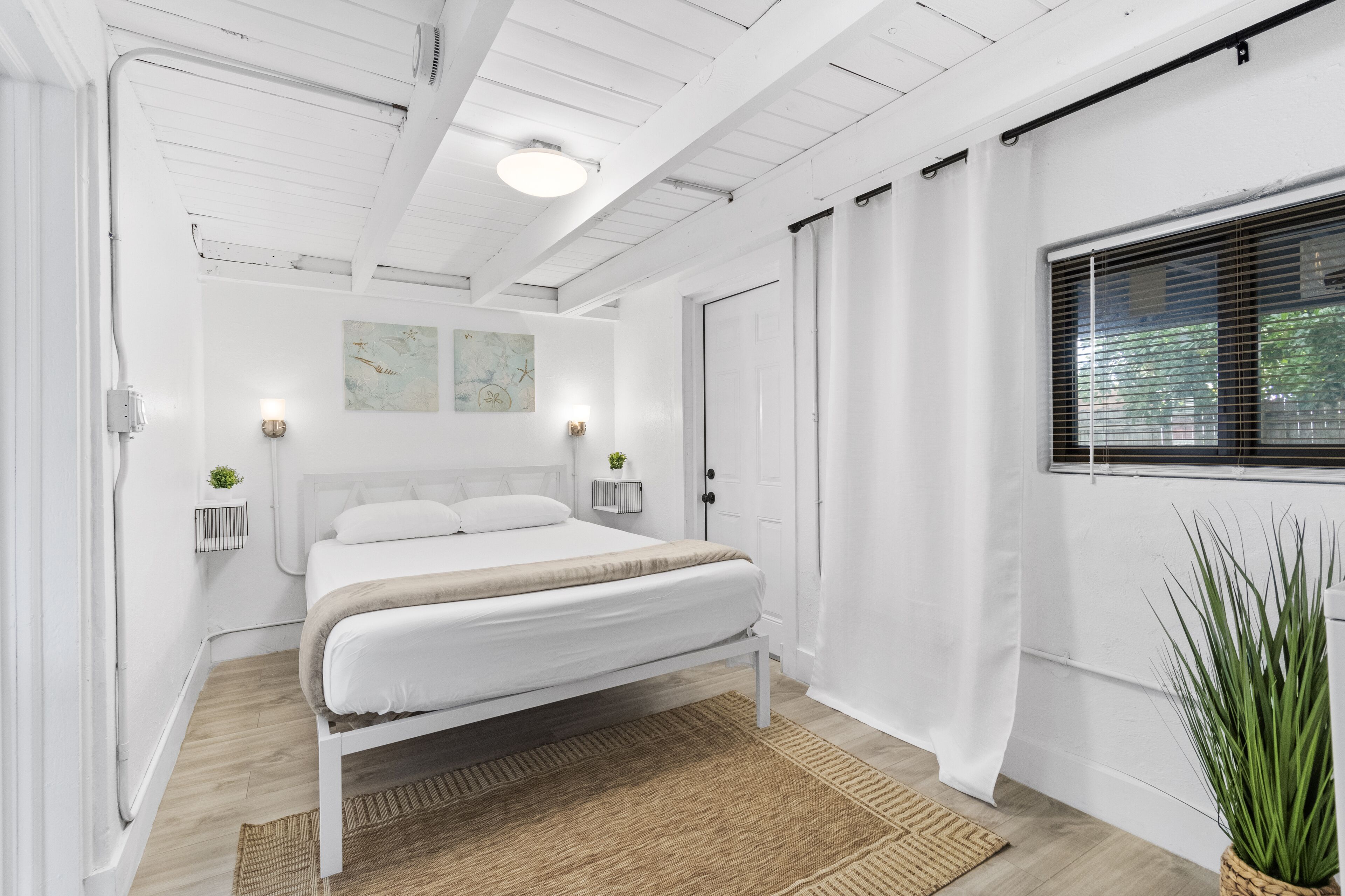 A bedroom with a bed, a window with blinds, a door, and a wall-mounted light fixture.