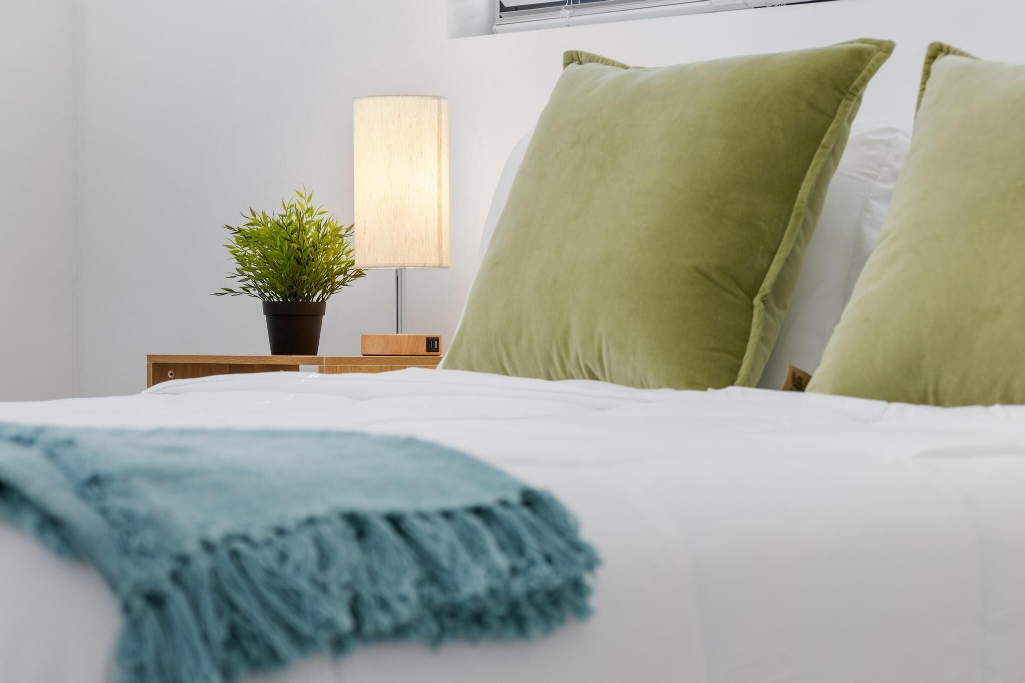 Una cama bien hecha con almohadas verdes, un plaid azul, una planta en la mesita de noche y una lámpara.