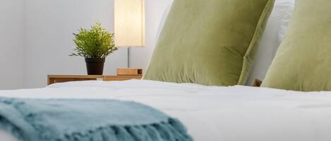 Una cama bien hecha con almohadas verdes, un plaid azul, una planta en la mesita de noche y una lámpara.