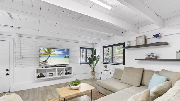 A modern living room with a white brick wall, a large flat-screen TV, a beige sofa, a wooden coffee table, and a potted plant. - Oasis by Sun Getaway Rentals Fort Lauderdale (Fort Lauderdale)