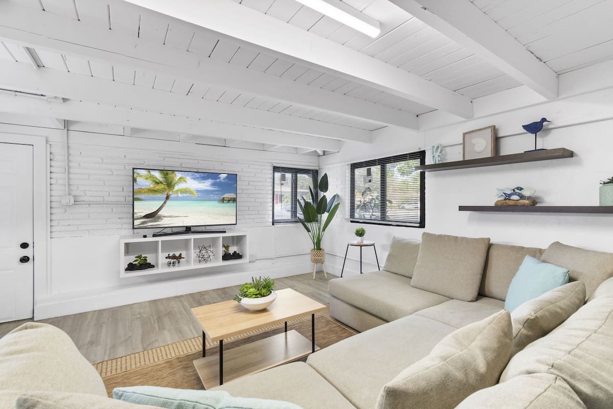 Een moderne woonkamer met een witte baksteenmuur, een grote flatscreen televisie, een beige bank, een houten salontafel en een potplant.