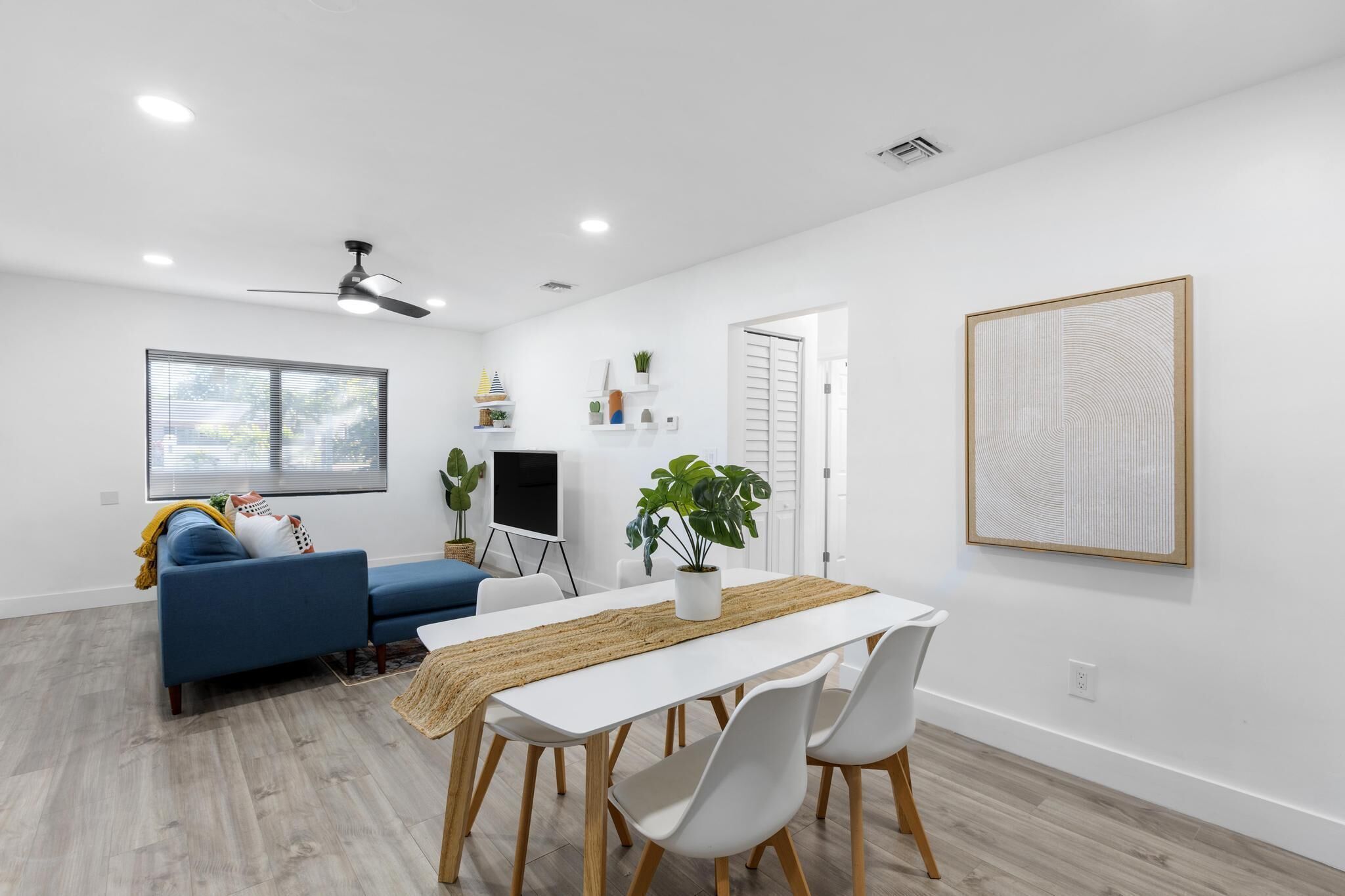 Uma sala de estar moderna com um sofá branco, uma mesa de jantar com cadeiras, um ventilador de teto e uma janela grande com persianas.