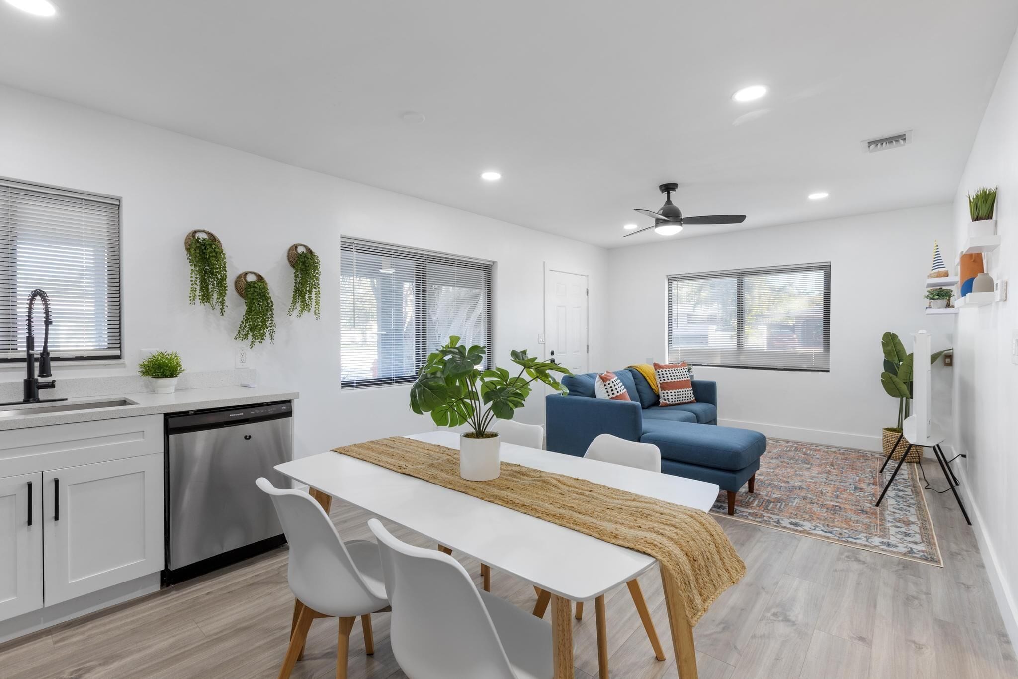 Una cocina moderna con armarios blancos, lavavajillas y un comedor con mesa y sillas blancas. Hay plantas colgantes, un sofá azul y un ventilador de techo en la sala de estar.