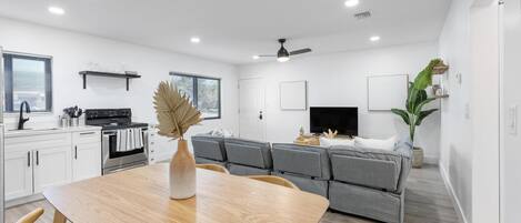 A modern kitchen with white cabinets, a dining table with chairs, a gray sofa, and a ceiling fan.