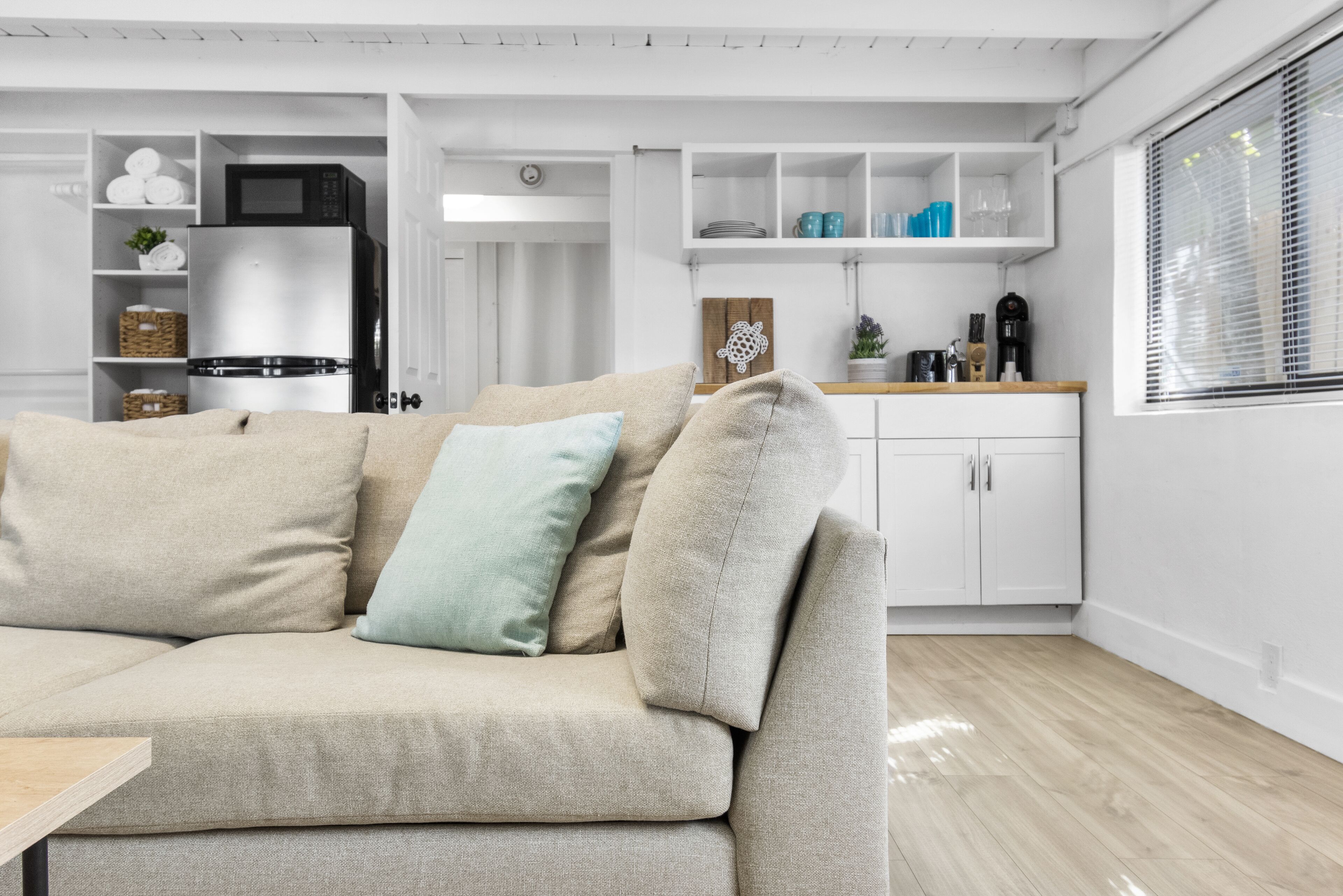 A modern living room with a beige sectional sofa, white cabinetry, and a kitchen area in the background.