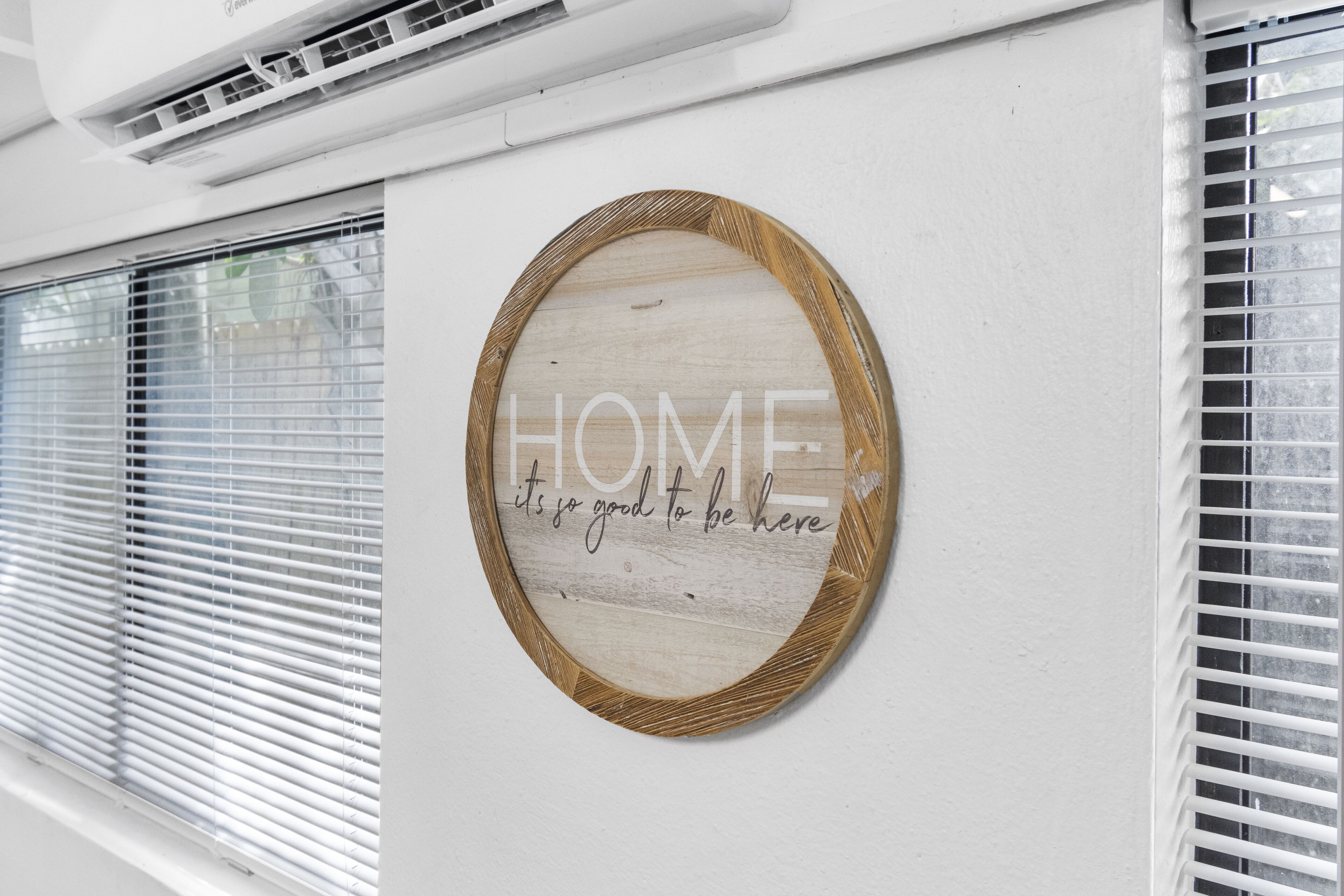 Un letrero de madera circular con la palabra 'HOME' y la frase 'it's so good to be here' sobre una pared blanca.