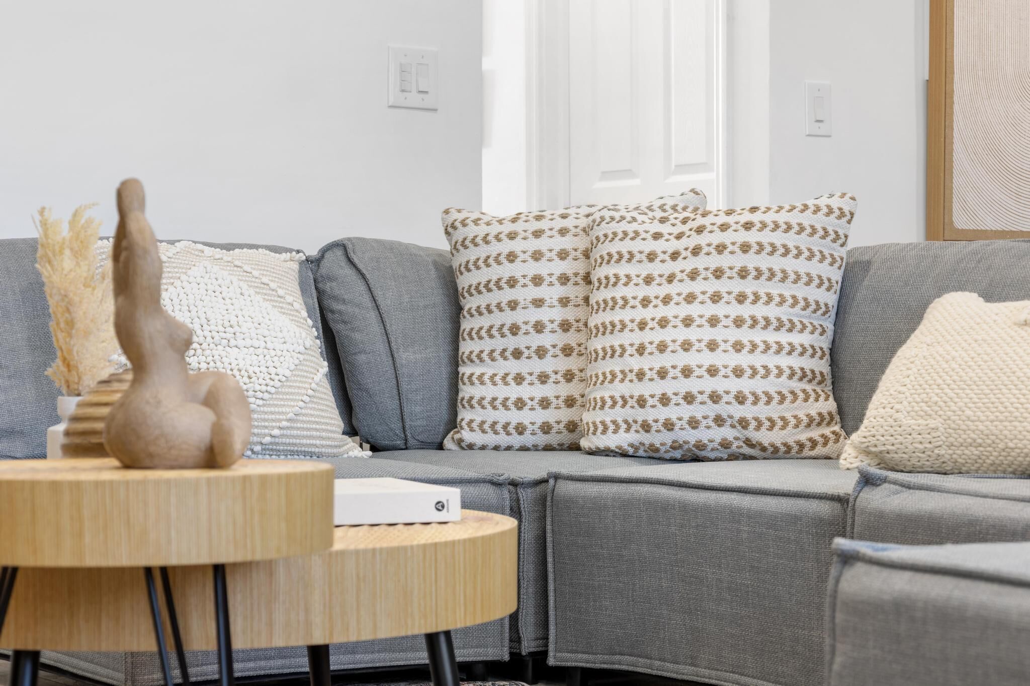 A modern living room with a grey sofa, patterned cushions, a small wooden table, and a white wall.