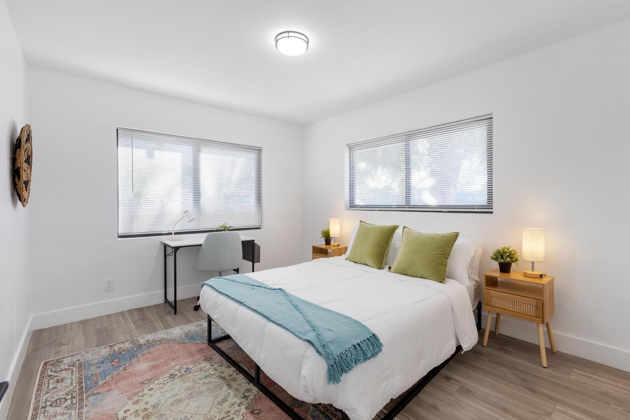 A bedroom with a bed, two bedside tables with lamps, a desk with a chair, and a large window with blinds.