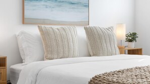 A neatly made bed with white bedding, two textured pillows, a framed ocean view painting, a bedside lamp, and a small plant on a wooden nightstand. - Oasis by Sun Getaway Rentals Fort Lauderdale (Fort Lauderdale)