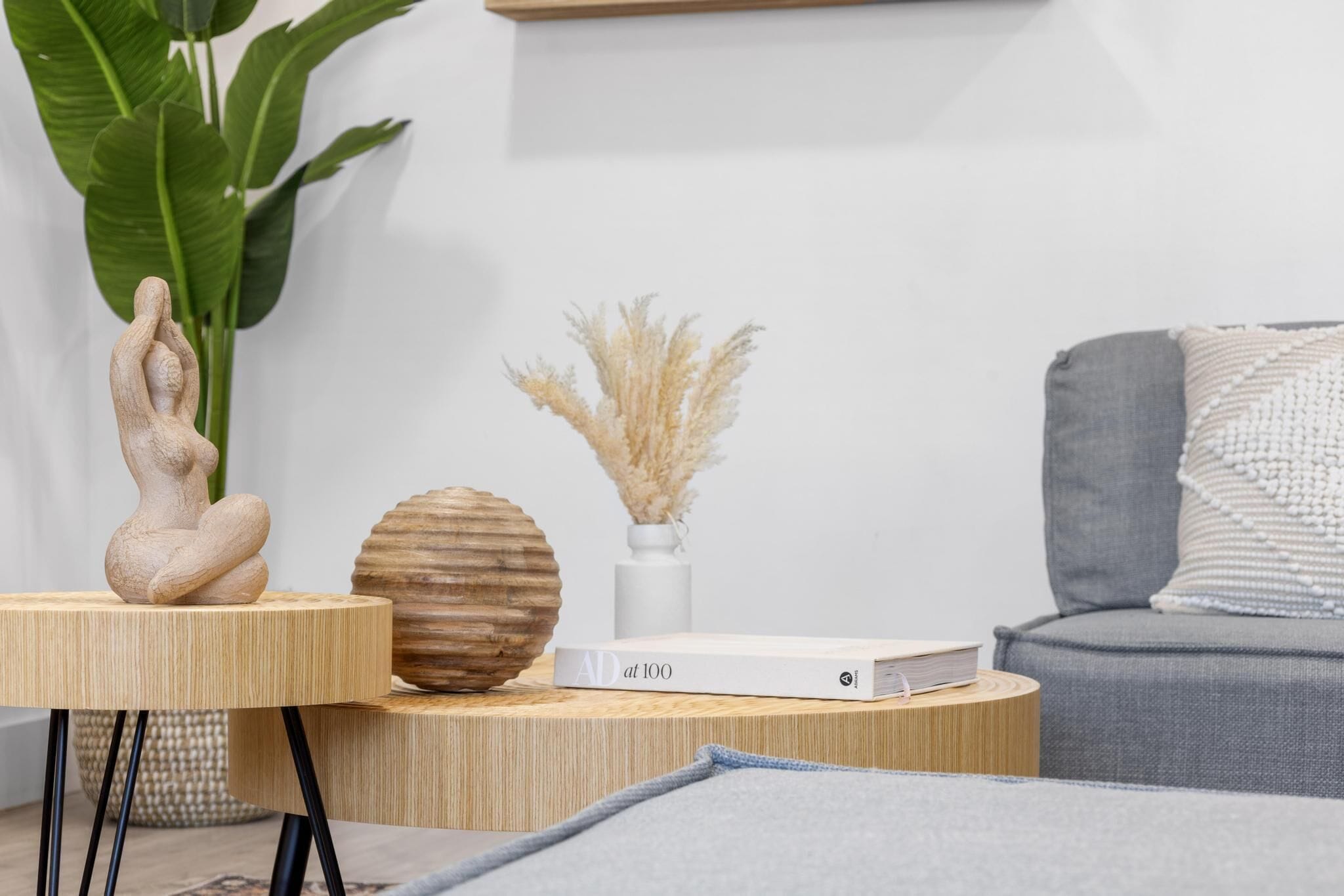 A modern living room with a wooden side table, a book titled 'AD at 100', a vase with pampas grass, and a grey sofa.
