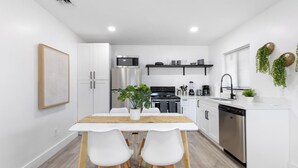 A modern kitchen with white cabinetry, a stainless steel refrigerator, a microwave, and a dishwasher. There is a wooden dining table with two chairs and a potted plant. - Oasis by Sun Getaway Rentals Fort Lauderdale (Fort Lauderdale)