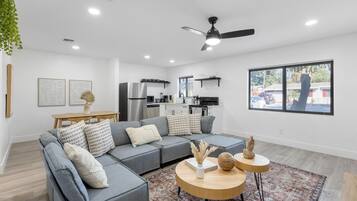 A modern living room with a grey sectional sofa, a round wooden coffee table, and a view of the kitchen in the background.