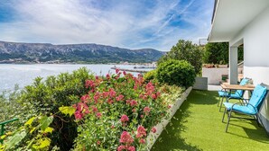 Un balcon avec vue sur la mer et les montagnes, un petit jardin agrémenté de fleurs roses et un coin salon avec des chaises bleues.