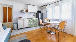 Une pièce compacte et bien éclairée, dotée d’un coin cuisine, d’un espace repas avec une table ronde et des chaises, d’un lit recouvert d’une couverture bleue et d’une fenêtre ornée de rideaux légers.