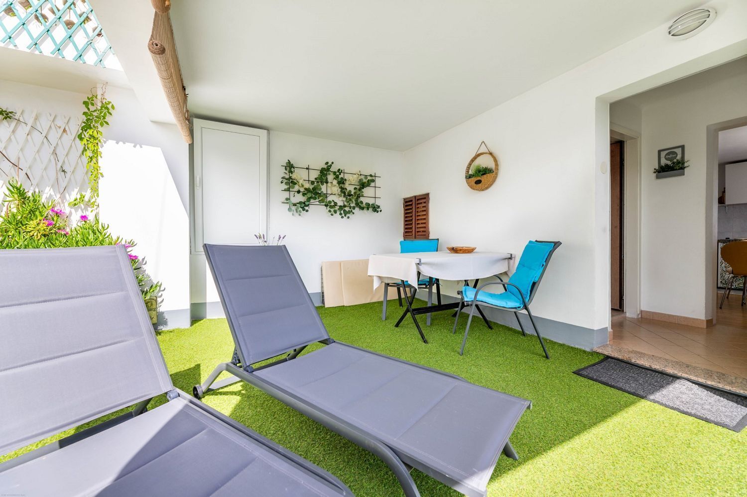 A sunlit patio with a table and chairs, surrounded by artificial grass and a white wall.