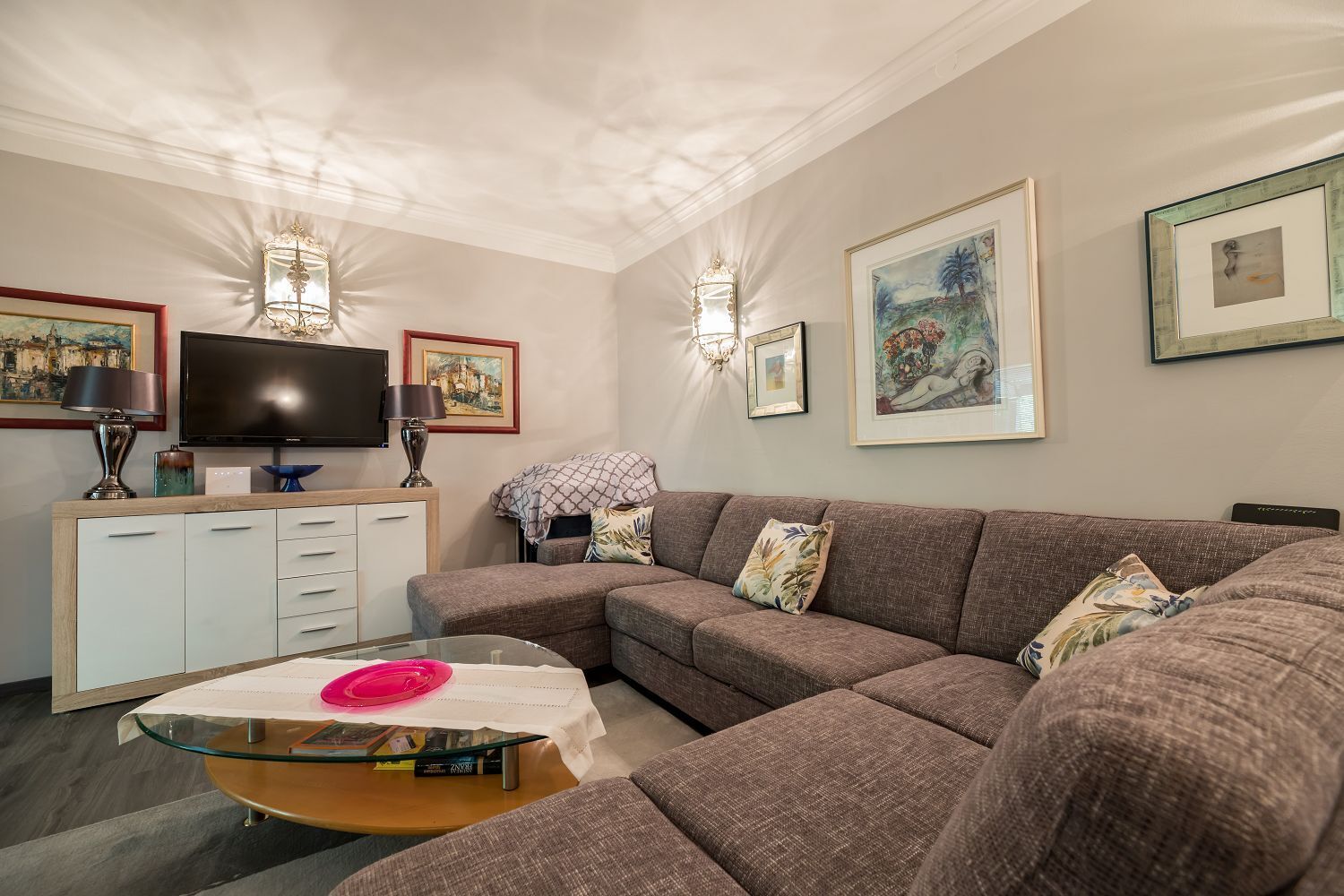 A living room with a sectional sofa, a glass coffee table, a flat-screen TV, a cabinet with drawers, and wall-mounted lights.